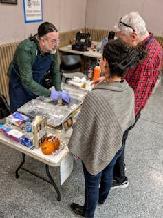 Man wearing rubber gloves hunches over at a table as he speaks with an adult woman and an adult man.